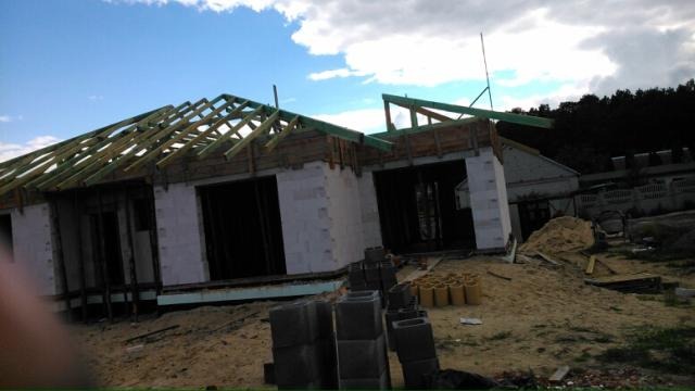 Budowa domu jednorodzinnego w stanie surowym otwartym, widoczne bloczki betonowe, drewniana konstrukcja dachu i niebo z chmurami.