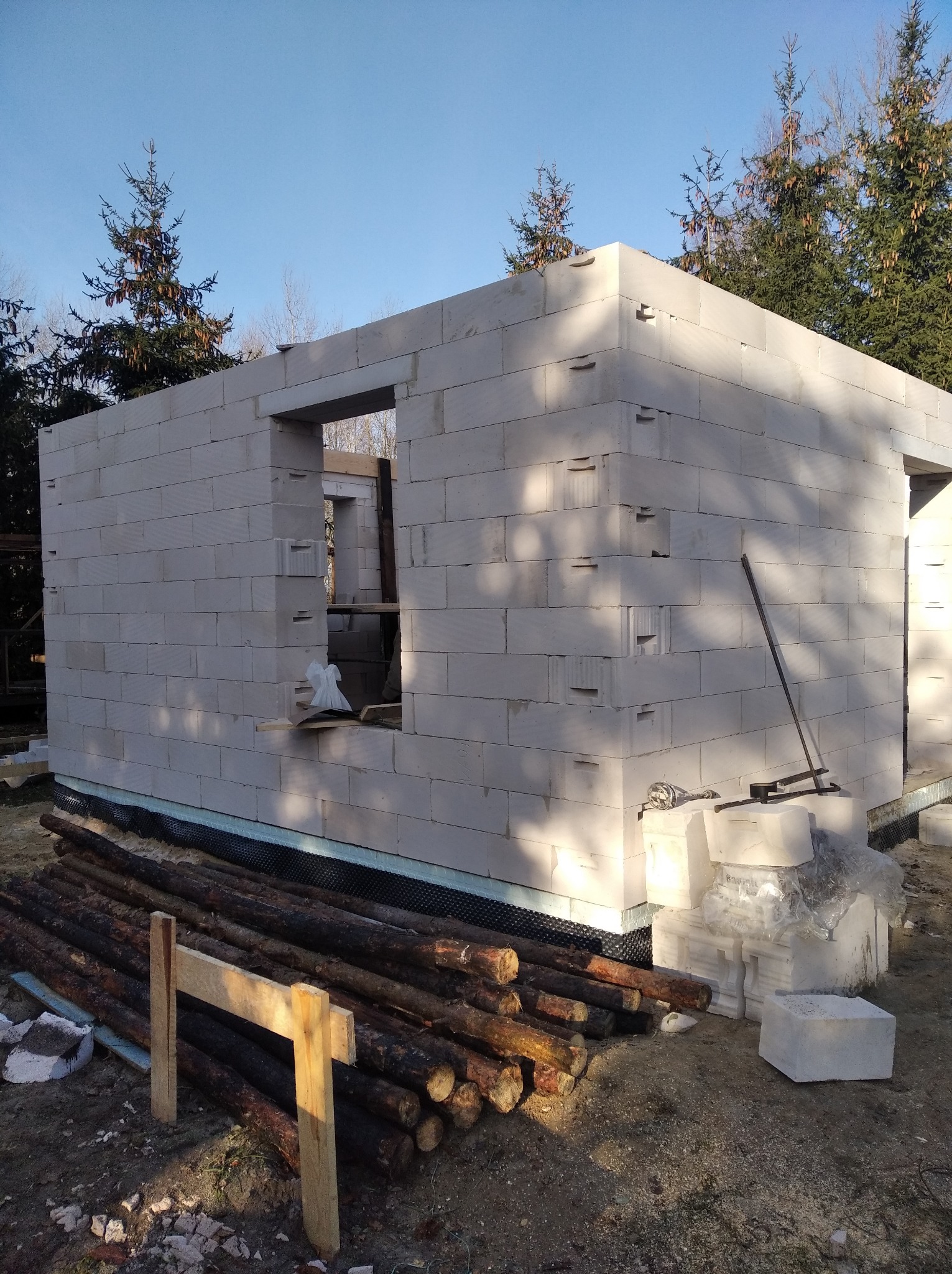 Wznoszenie ścian domu z bloczków betonu komórkowego, widoczne otwory okienne i drzwiowe, przygotowane drewno i narzędzia budowlane na placu budowy.