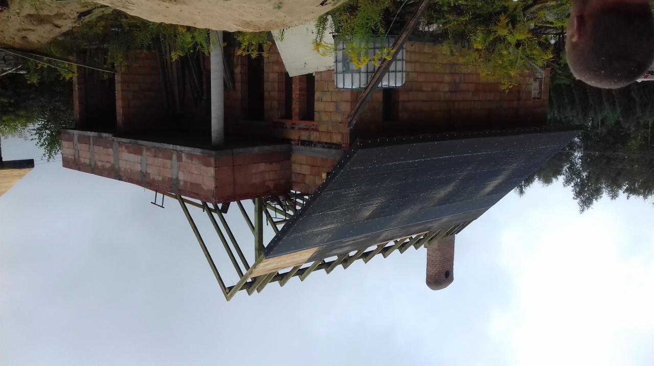 Budowa domu z cegły: widoczna konstrukcja dachu pokryta czarną membraną, komin z cegły, ściany w trakcie wznoszenia, częściowo zbrojone, drewniane elementy konstrukcyjne. W tle zieleń.