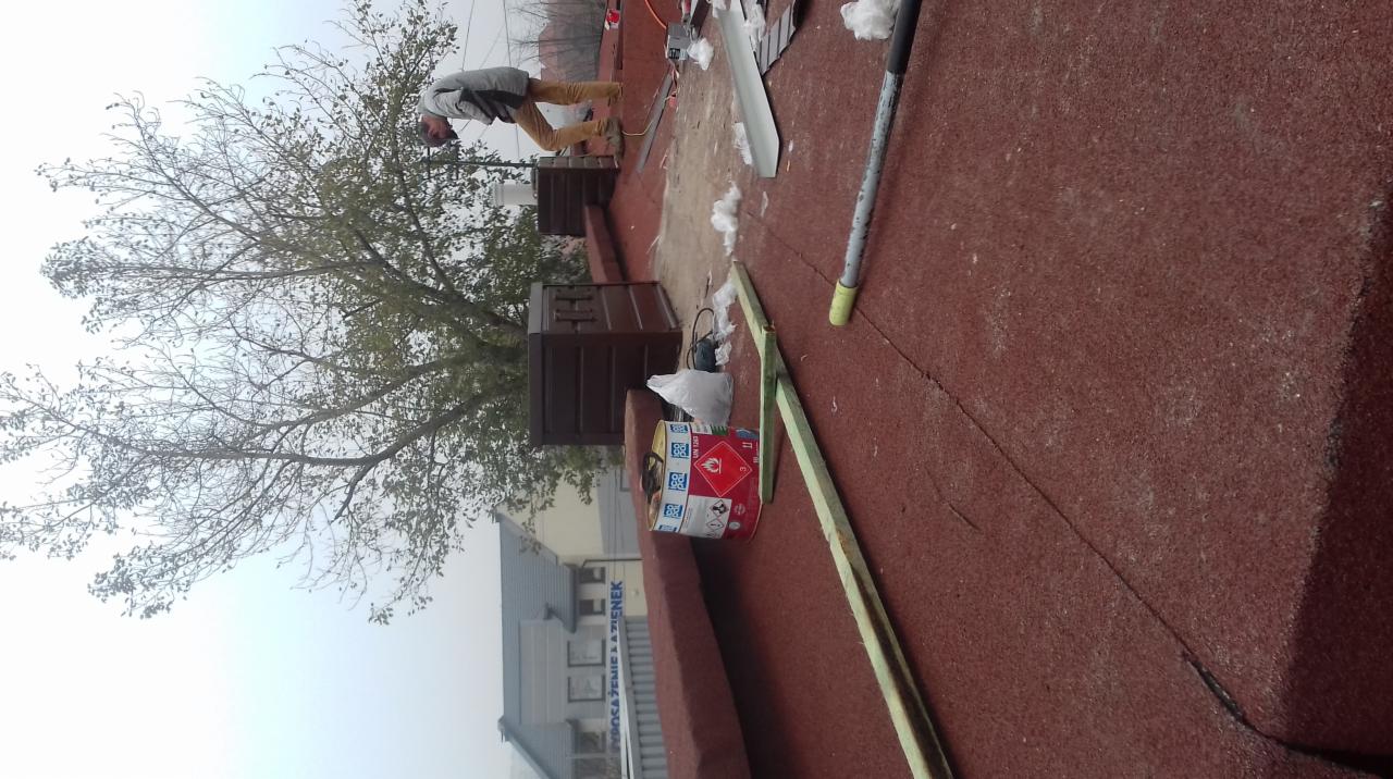 Pracownik w trakcie pracy na dachu pokrytym czerwoną papą, widoczne kominki wentylacyjne, puszka z klejem i narzędzia pracy.