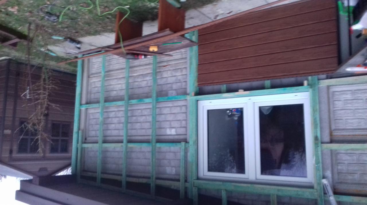 Montaż brązowej podbitki dachowej na drewnianym ruszcie, widoczne okno dachowe i fragment elewacji budynku obok.