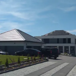 Nowoczesny dom jednorodzinny z panelami słonecznymi na dachu, widok z poziomu podjazdu z kostki, kilka samochodów zaparkowanych na posesji.