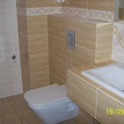 Wykończona łazienka z beżowymi płytkami imitującymi drewno, podwieszana toaleta, umywalka z baterią i dekoracyjnym pasem płytek z motywem fal.