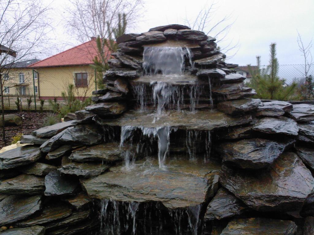 Kaskada wodna z ciemnego kamienia w ogrodzie, z widocznym domem w tle i młodymi drzewkami iglastymi.