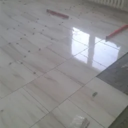 Podłoga w trakcie układania płytek ceramicznych z użyciem krzyżyków dystansowych i poziomicy; widoczne odbicie okna w polerowanej powierzchni płytek.