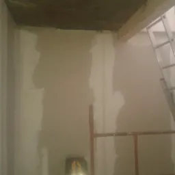 Przygotowanie ściany do malowania: częściowo zagruntowana ściana z widocznym laserowym niwelatorem, fragment rusztowania i drabiny w tle.