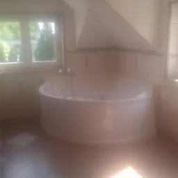 Jasna łazienka na poddaszu z narożną wanną z hydromasażem, wykończona kafelkami z delikatnym wzorem, okno z widokiem na zieleń i punktowe oświetlenie sufitowe.