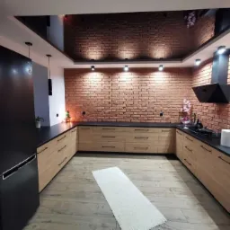 Nowoczesna kuchnia z ceglaną ścianą, drewnianymi szafkami z czarnymi uchwytami, czarnym blatem i podwieszanym sufitem z lustrzanym wykończeniem. Na podłodze jasne panele i biały dywanik.