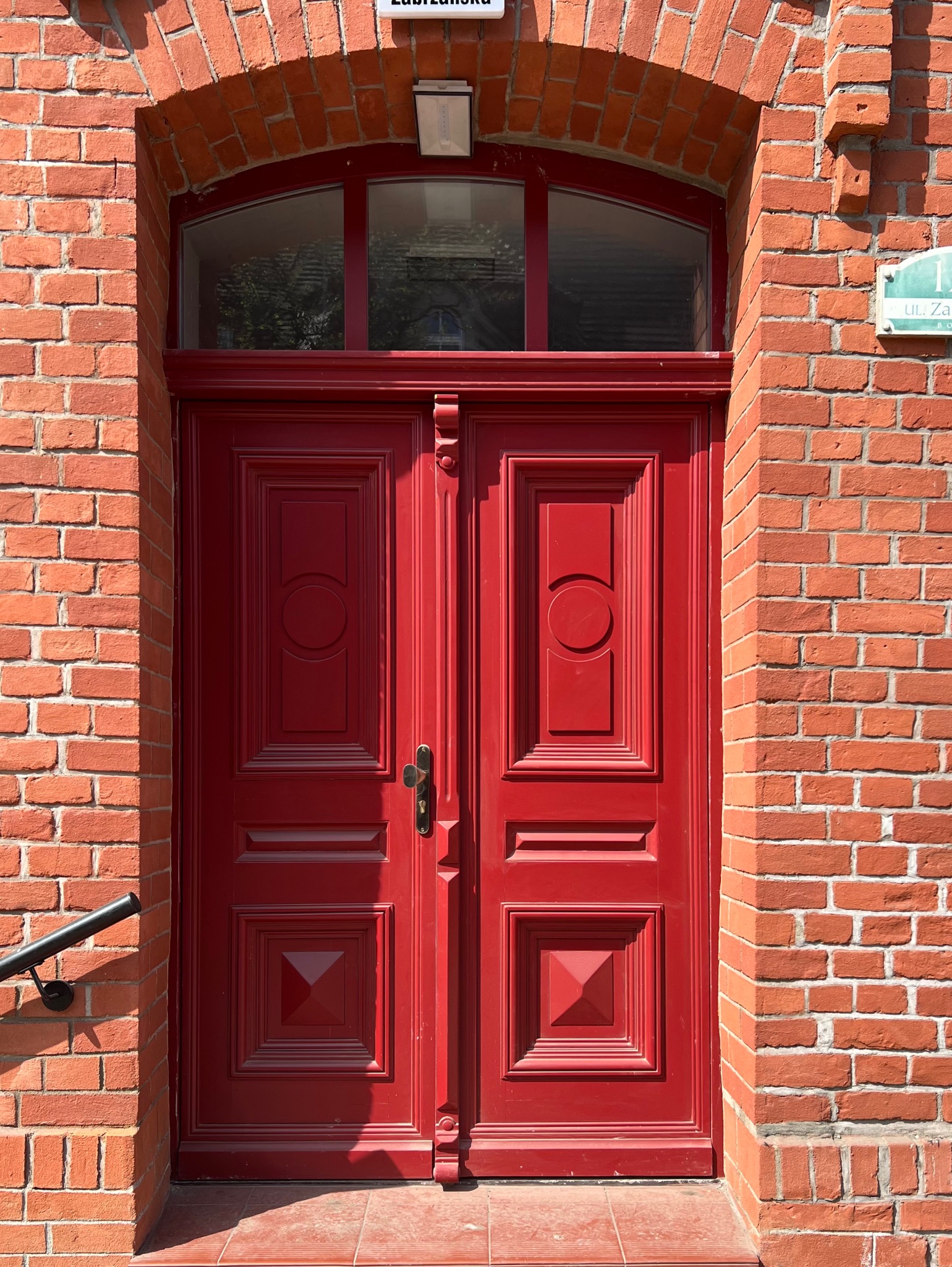 Czerwone, drewniane drzwi wejściowe z dekoracyjnymi panelami, umieszczone w ceglanej fasadzie budynku z widocznym fragmentem ulicy i tabliczką z nazwą ulicy.