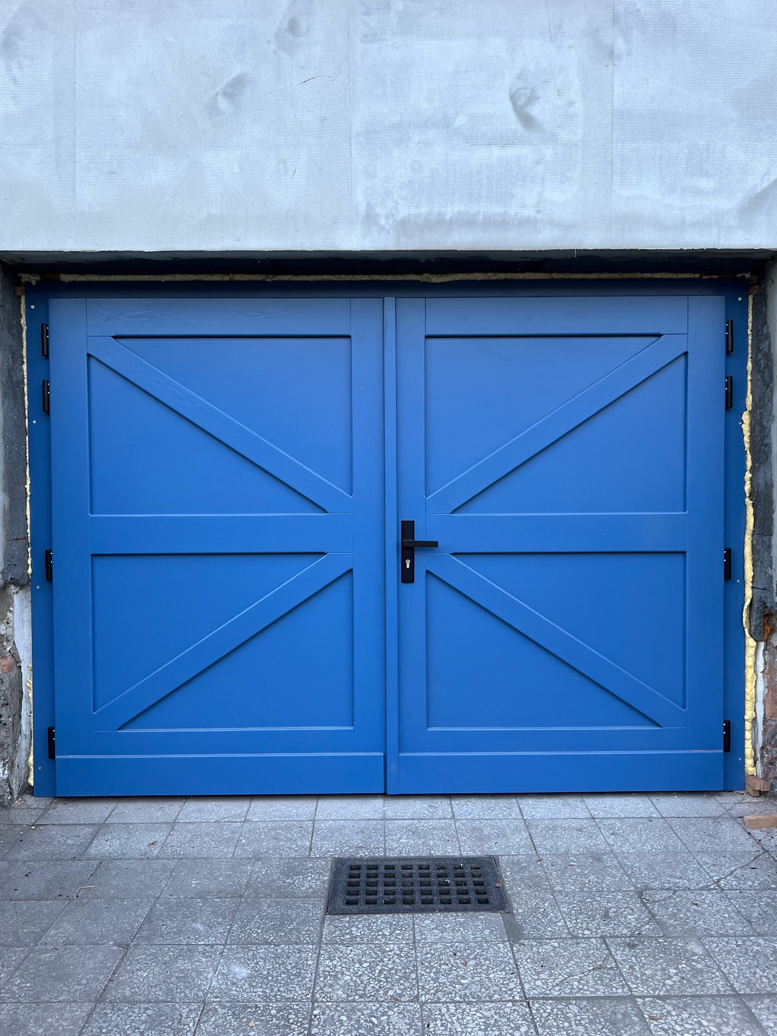 Dwuskrzydłowe, niebieskie drzwi z ukośnymi listwami w kształcie litery X, czarną klamką i widoczną pianką montażową w szparach między ościeżnicą a murem.