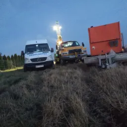 Żółty samochód ciężarowy z wiertnicą i biały van firmy Geoplantex na zarośniętym polu o zmierzchu.