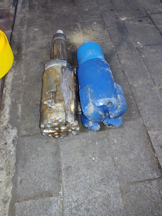 Dwie głowice wiertnicze leżące na szarych płytkach chodnikowych: jedna w kolorze srebrnym, z widocznymi śladami użytkowania i licznymi małymi wypustkami, a druga pomalowana na niebiesko z trzema...