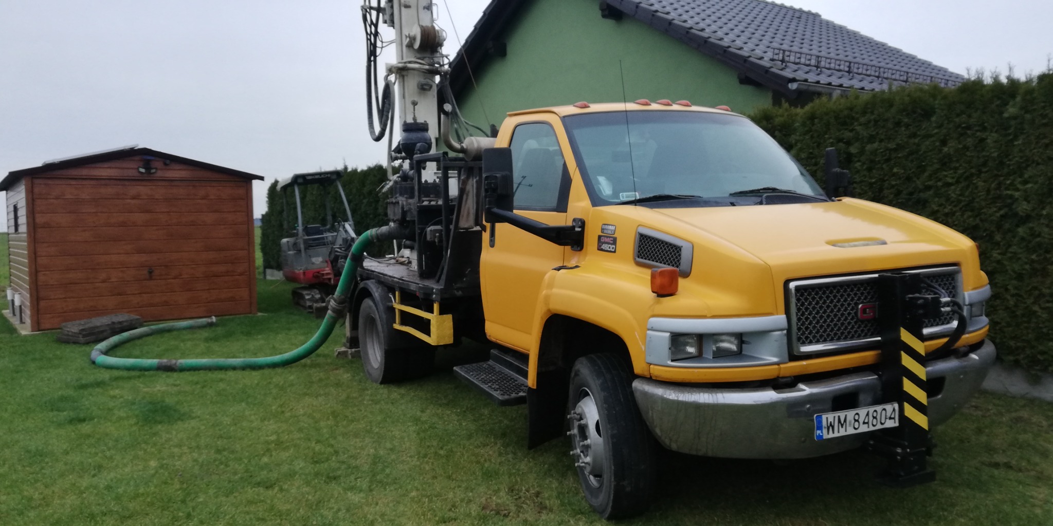 Żółta ciężarówka GMC 4500 z zamontowaną wiertnicą do studni na trawniku, w tle widoczny mały domek gospodarczy i minikoparka.