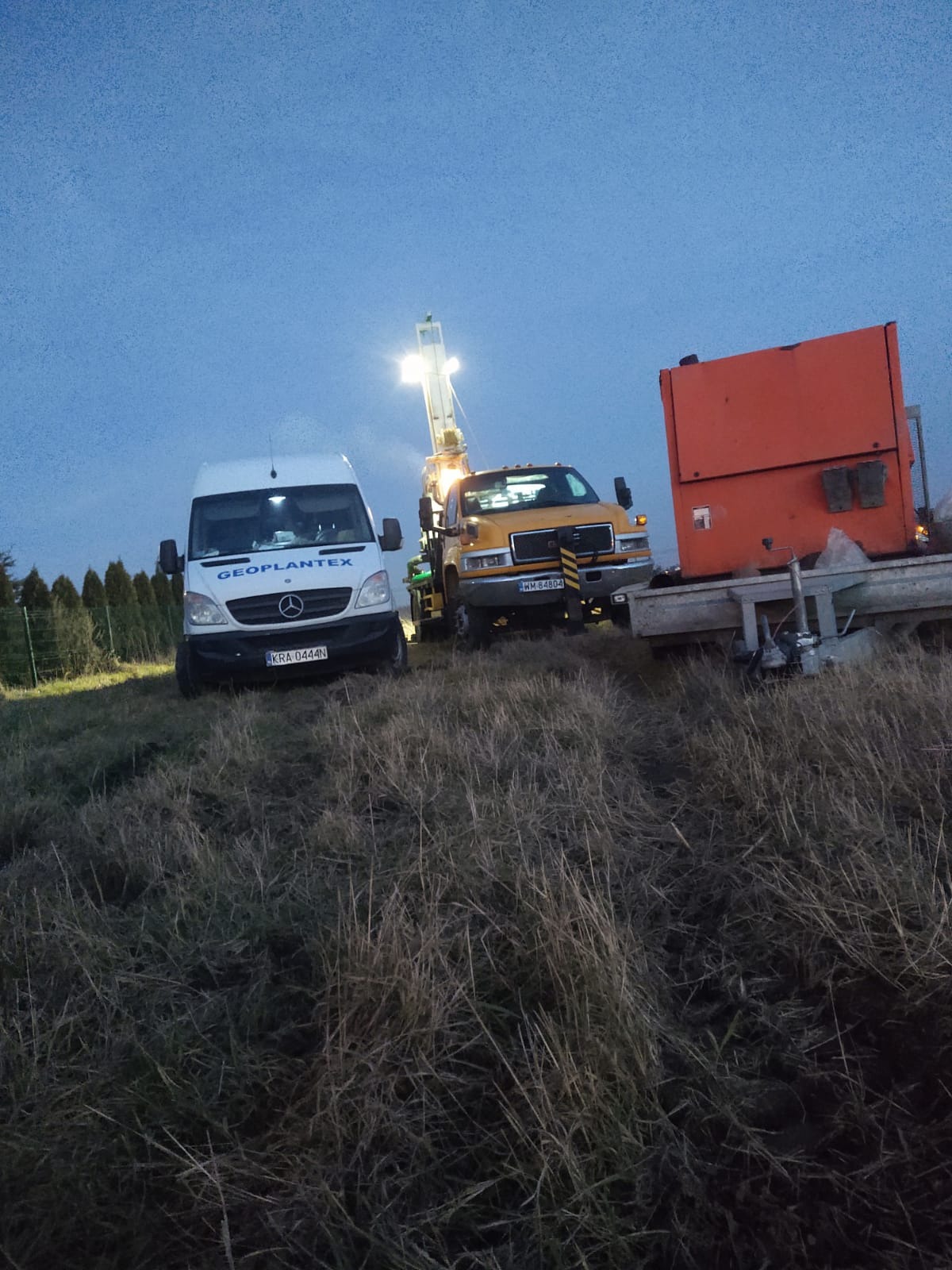 Żółty samochód ciężarowy z wiertnicą i biały van firmy Geoplantex na zarośniętym polu o zmierzchu.
