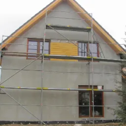 Dom w trakcie ocieplania: elewacja pokryta szarym tynkiem, rusztowanie przy ścianie, okna z brązowymi ramami, zabezpieczone deskami otwory okienne na górze, widoczna konstrukcja dachu z drewnianymi...