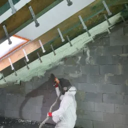 Pracownik w kombinezonie ochronnym aplikuje piankę izolacyjną na poddaszu z widocznymi krokwiami i metalowymi uchwytami montażowymi.