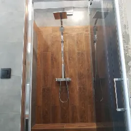 Wykończona kabina prysznicowa z drewnopodobnymi panelami na ścianie i deszczownicą, widoczny fragment ściany z szarymi płytkami oraz czarny włącznik światła.