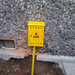 Żółta skrzynka gazowa z symbolem płomienia i napisem GAZ, umieszczona na żółtej rurze, na tle ściany z kamieni i wykopu.