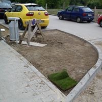 Przygotowany teren pod trawnik z rolki, widoczny fragment rozwiniętej rolki trawy oraz zwinięta rolka w rogu. W tle zaparkowane samochody.