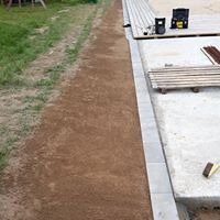 Ułożone obrzeża betonowe oddzielające trawnik od tarasu, przygotowane pod montaż automatycznego systemu koszenia. Widoczny fragment tarasu, trawnika i narzędzia pracy.