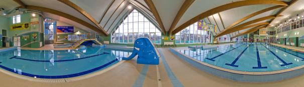 Panorama krytego basenu z niebieskim słoniem-zjeżdżalnią, drewnianą konstrukcją dachu i oknami sięgającymi sufitu.