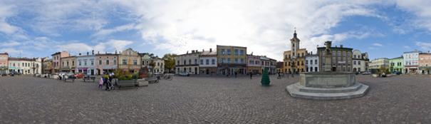 Panoramiczny widok na rynek miejski z fontanną, kolorowe kamienice i ratusz z wieżą zegarową w tle, wykonany z użyciem obiektywu szerokokątnego, ukazujący rozległą przestrzeń placu.
