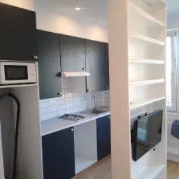 Nowoczesna, mała kuchnia z zabudową w odcieniach szarości i bieli, z wbudowaną płytą gazową i telewizorem w regale oddzielającym przestrzeń.