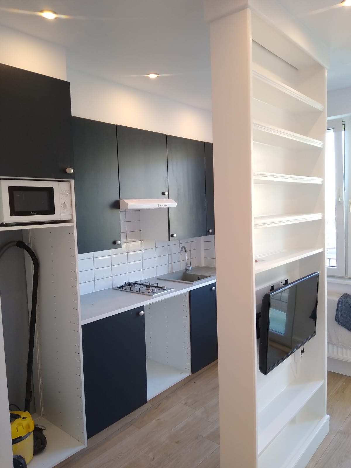 Nowoczesna, mała kuchnia z zabudową w odcieniach szarości i bieli, z wbudowaną płytą gazową i telewizorem w regale oddzielającym przestrzeń.