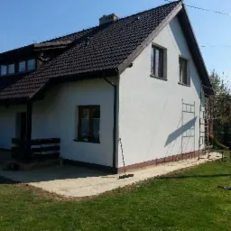 Dom jednorodzinny z ciemnym dachem z dachówki, białą elewacją i rusztowaniem przy ścianie. Widoczny fragment trawnika i betonowego podjazdu.