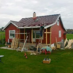 Czerwony dom z brązowym dachem w trakcie budowy werandy. Pracownik na drabinie montuje konstrukcję. Na trawniku leżą deski i narzędzia.