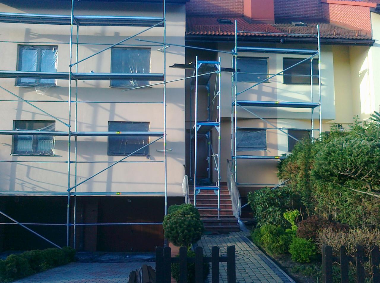 Elewacja budynku w trakcie prac remontowych, zabezpieczone okna folią, rusztowania, chodnik z kostki, krzewy. Widok na dwukondygnacyjny budynek.