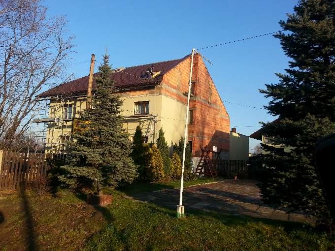 Dom w trakcie renowacji dachu z widocznymi rusztowaniami, częściowo odsłoniętą konstrukcją i ekipą na dachu. Na pierwszym planie iglaki i trawnik.