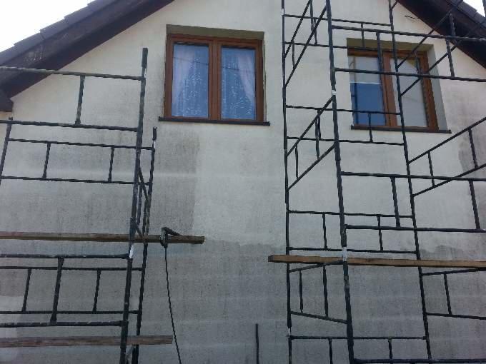 Elewacja domu w trakcie czyszczenia, widoczne rusztowania i fragmenty oczyszczonej ściany. Dwa okna z brązowymi ramami na tle jasnej elewacji.