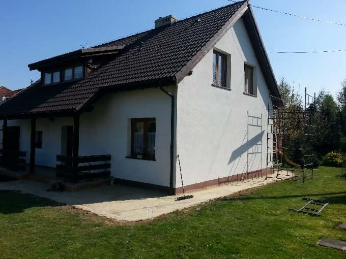 Dom jednorodzinny z ciemnym dachem z dachówki, białą elewacją i rusztowaniem przy ścianie. Widoczny fragment trawnika i betonowego podjazdu.