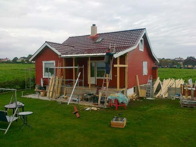 Czerwony dom z brązowym dachem w trakcie budowy werandy. Pracownik na drabinie montuje konstrukcję. Na trawniku leżą deski i narzędzia.