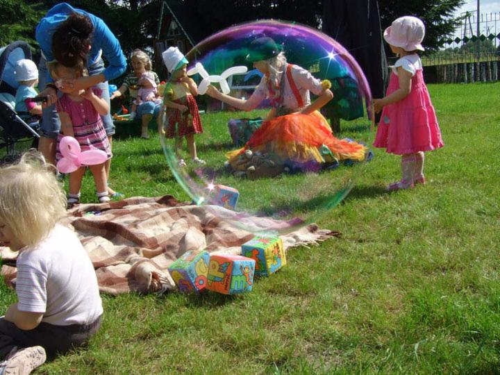 Animator w stroju klauna dmucha ogromne bańki mydlane dla dzieci na trawie, w tle inne dzieci i rodzice.