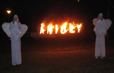 Dwie osoby przebrane za anioły na szczudłach stoją po bokach płonącego napisu, widoczne w nocy.