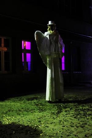 Osoba przebrana za anioła na szczudłach, w białym stroju i kapeluszu, stoi na zewnątrz budynku z oknami oświetlonymi na fioletowo, na ziemi leżą konfetti.