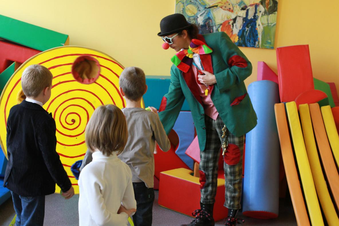 Klaun w zielonej marynarce i kraciaste spodnie z dzwoneczkami, rozmawia z grupą dzieci na tle kolorowych piankowych kształtów i abstrakcyjnego obrazu.
