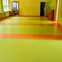 Wnętrze pokoju z podłogą z wykładziny w odcieniach zieleni i pomarańczy, widoczne okna z grzejnikami, drzwi do kolejnych pomieszczeń.