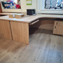 Biurko narożne z jasnego drewna z szufladami i szafką, na blacie drukarka i dwa podwyższenia, w tle okno z stertą dokumentów.