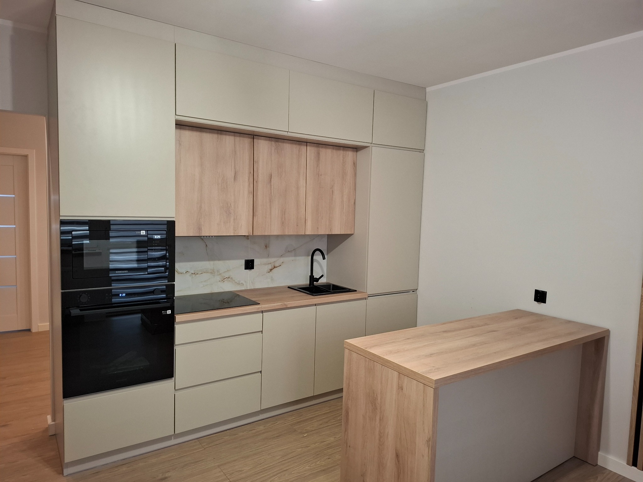 Nowoczesna kuchnia z beżowymi szafkami i drewnianymi akcentami, czarny zlew i bateria na tle marmurowej ściany, sprzęt AGD w zabudowie, wyspa kuchenna z blatem z drewna.