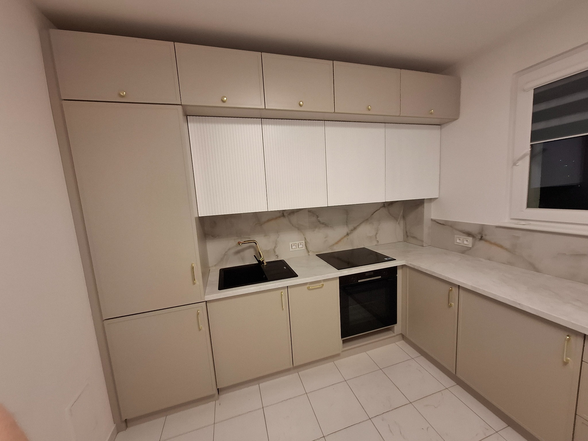 Jasna kuchnia z beżowymi szafkami, złotymi uchwytami, białymi frontami górnych szafek z pionowymi żłobieniami i blatem imitującym marmur.