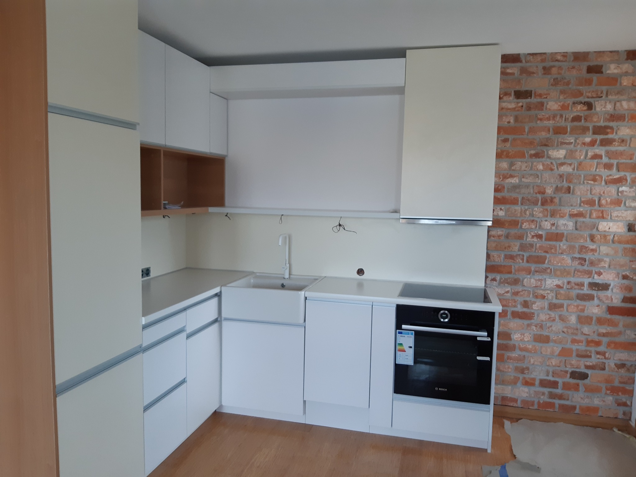 Jasna, minimalistyczna kuchnia z białymi frontami, ceramicznym zlewem i ścianą z czerwonej cegły w tle, widoczne niedokończone podłączenia elektryczne.