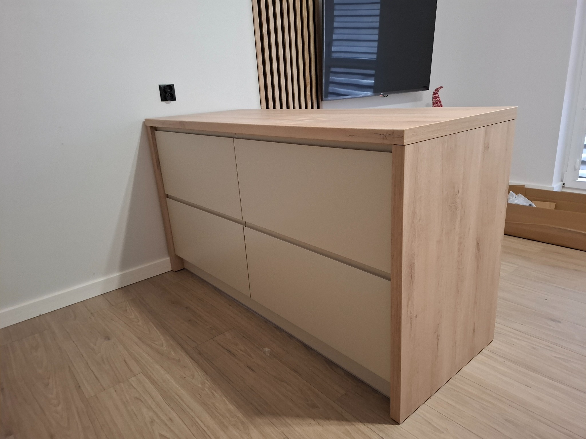 Niska komoda z czterema szufladami w odcieniach beżu i jasnego drewna, z blatem z litego drewna, umieszczona w nowoczesnym wnętrzu z drewnianą ścianką dekoracyjną i telewizorem w tle.