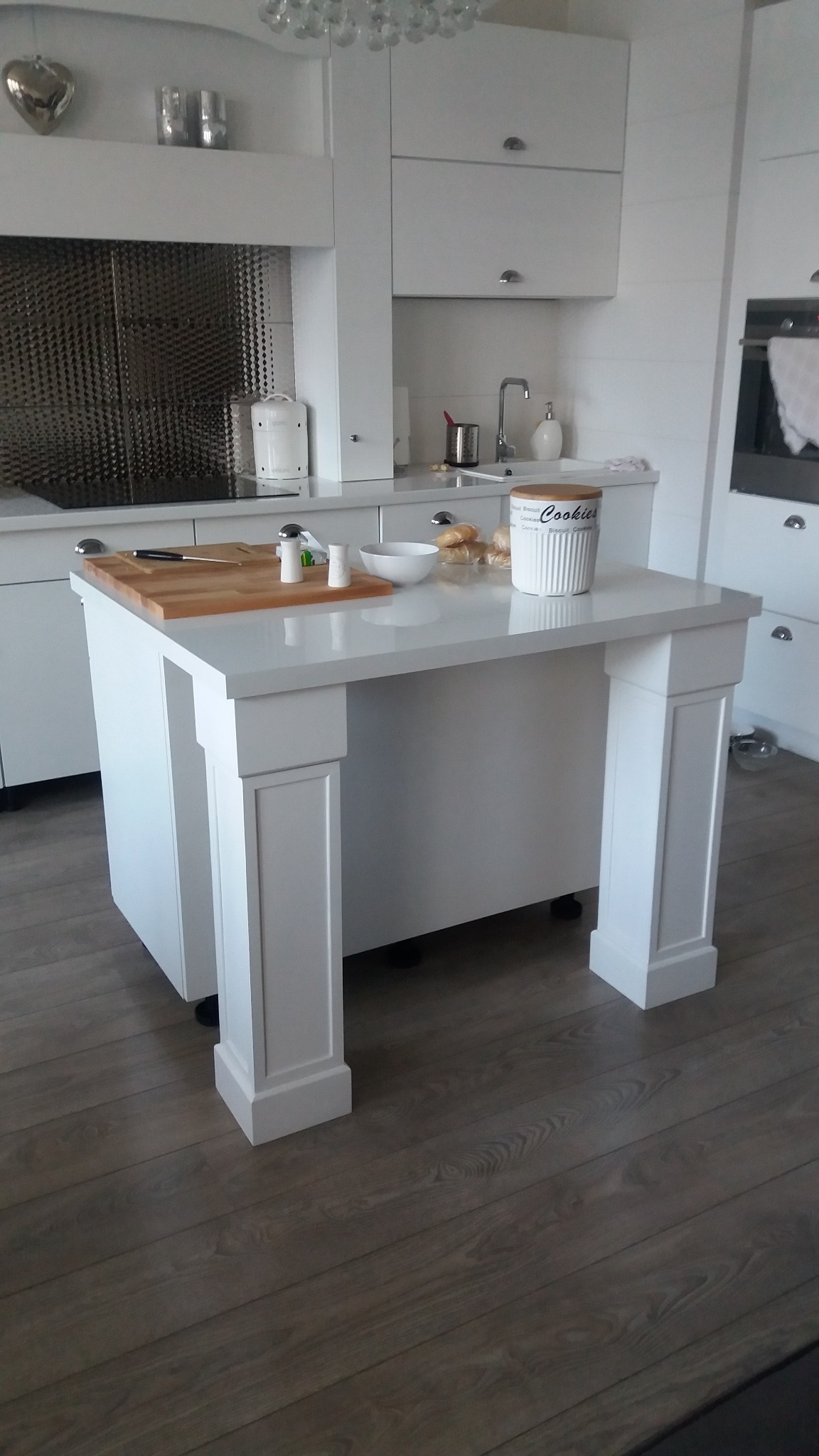Biała wyspa kuchenna z blatem, deska do krojenia, przyprawy, miska i pojemnik na ciasteczka, na tle białych szafek kuchennych i drewnianej podłogi.