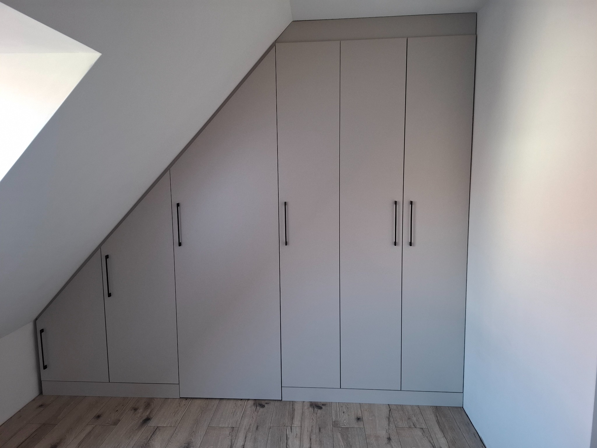Szafa wnękowa z szarymi frontami i czarnymi, minimalistycznymi uchwytami, dopasowana do skosu poddasza, na drewnianej podłodze.