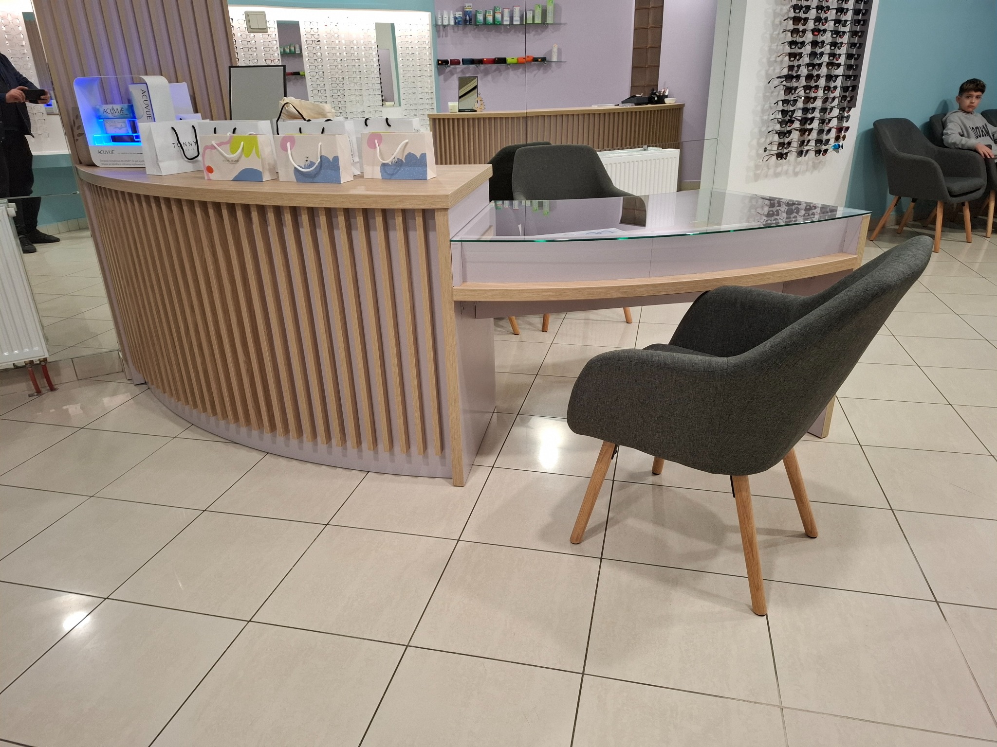 Recepcja w salonie optycznym z drewnianymi lamelami i szklanym blatem, obok szare krzesło na drewnianych nogach, w tle ekspozycja okularów