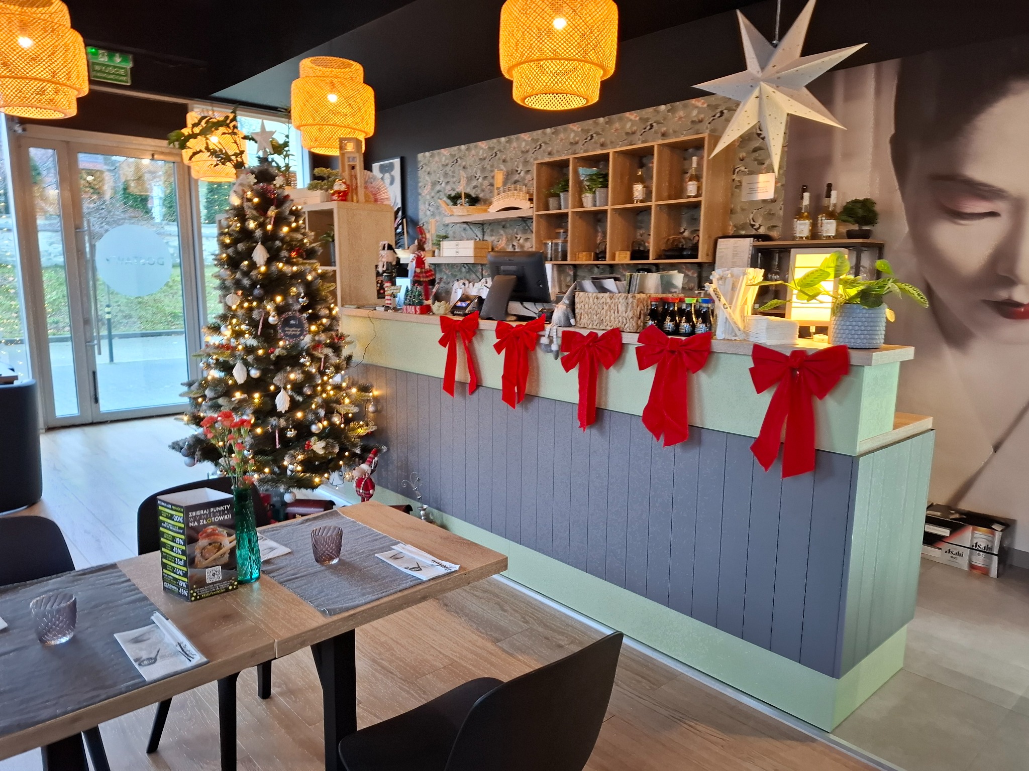 Wnętrze lokalu gastronomicznego z ladą recepcyjną udekorowaną na święta Bożego Narodzenia, widoczna choinka, drewniane stoły z nakryciami i krzesła, na ścianie mural z wizerunkiem kobiety.
