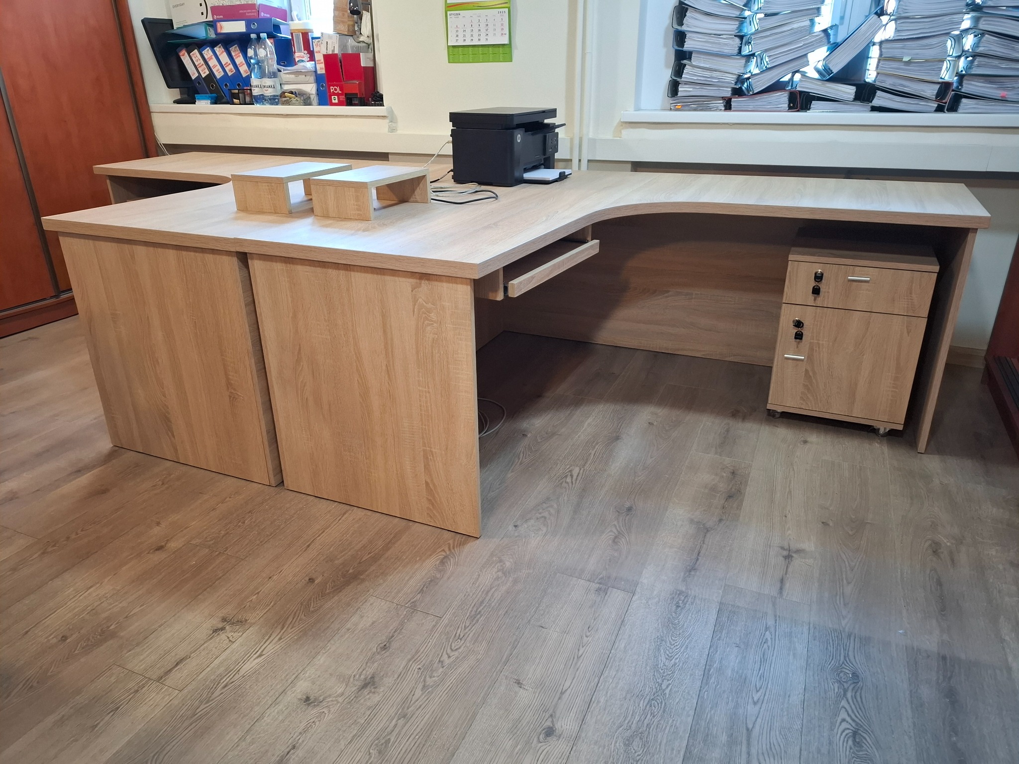 Biurko narożne z jasnego drewna z szufladami i szafką, na blacie drukarka i dwa podwyższenia, w tle okno z stertą dokumentów.